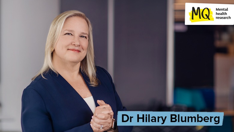 Dr Hilary Blumberg stands clasping her hands with a gentle smile looking to camera with blonde hair, navy blazer, white top and confident expression