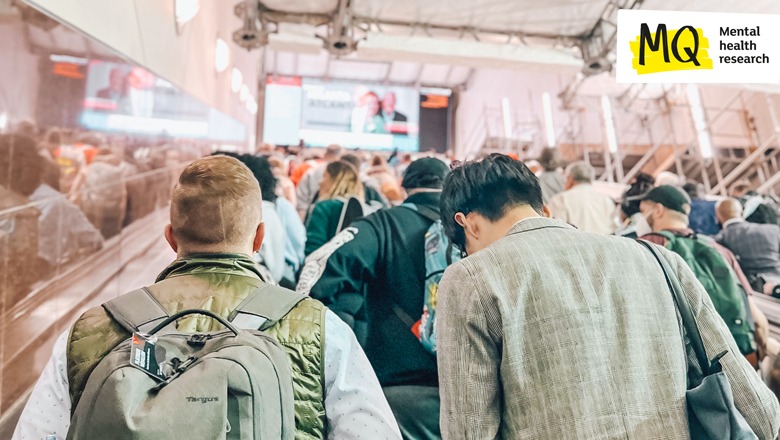 Commuters are crowded, we see their backs and backs of their heads in a busy public transport terminal. There is a bright digital screen far ahead of them.