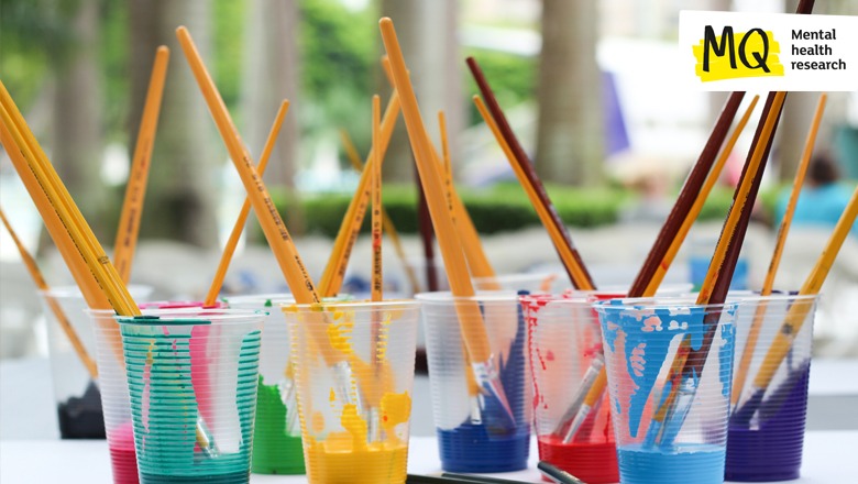 Colourful paints in plastic cups stand on a tabletop with paintbrushes sitting in them waiting to be used