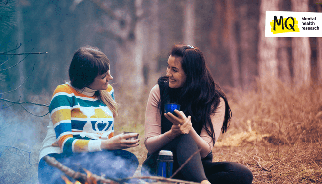 Two women are talking with smiles on their faces by a campfire in the woods