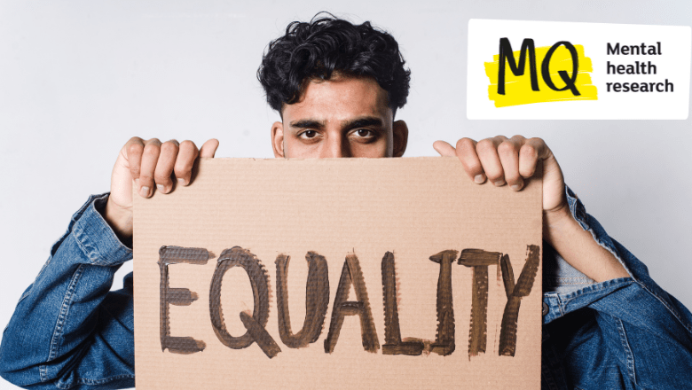 A male presenting figure holds a cardboard protest sign in front of his face, hiding his mouth and nose. The sign reads "Equality".