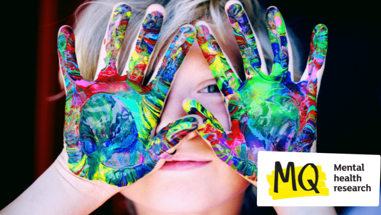 A young child holds their hands in front of their face. Their palms are covered with beautiful swirls of multicoloured paint