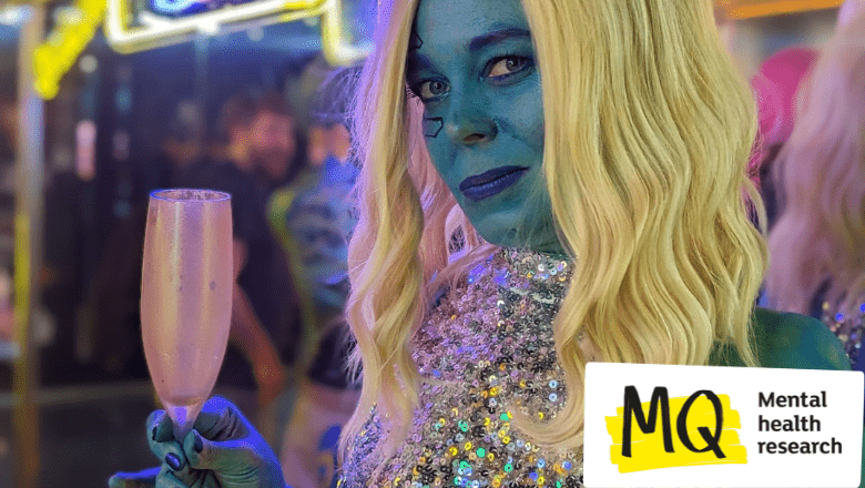 Painted blue, with blue make up, wearing a blonde wig and a glittery silver sequin dress, holding a gold champagne flute, a woman (Juliette Burton - the writer, cosplayer and comedian) smiles to camera.
