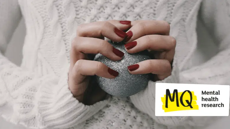 We can see a white woman's hands with dark red nail varnish on her nails clutching a glittery silver bauble. We also see she is wearing a soft white fluffy jumper. We do not see more than her torso, hands, wrists and sleeves. The colour wash is cold and her grip looks tense.