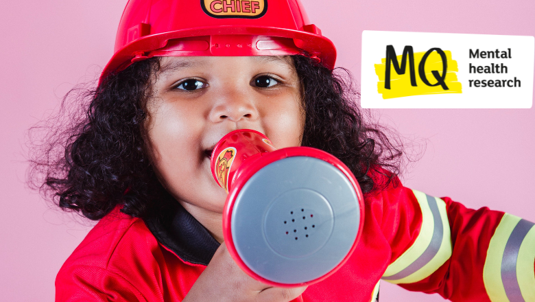A young children with dark curly hair wears a kids fireman costume with plastic helmet and holds a toy loudhailer into which she is shouting. The background behind her is pale pink.