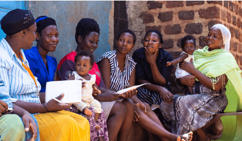 six Ugandan women sit outside talking with each other. two have babies